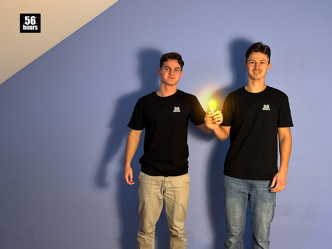 Team 56hours 2024 Die beiden Gründer von 56hours stehen vor einer blauen Wand und halten eine leuchtende Glühbirne.